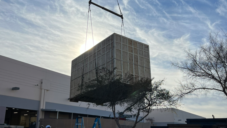 Oversized wood crate lifting construction equipment by crane, designed for rugged transport and protection in industrial environments.