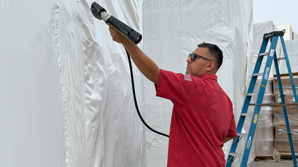 A Crating Technology technician applies heat-shrink overwrap to a wooden crate bound for an offshore oil rig, using a heat gun to ensure a moisture-tight, corrosion-resistant seal for marine transport.