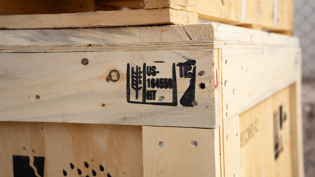 Close-up of a wooden shipping crate stamped with ISPM 15 compliance mark, showing wheat symbol, country code “US,” registration number, and heat treatment code “HT.”