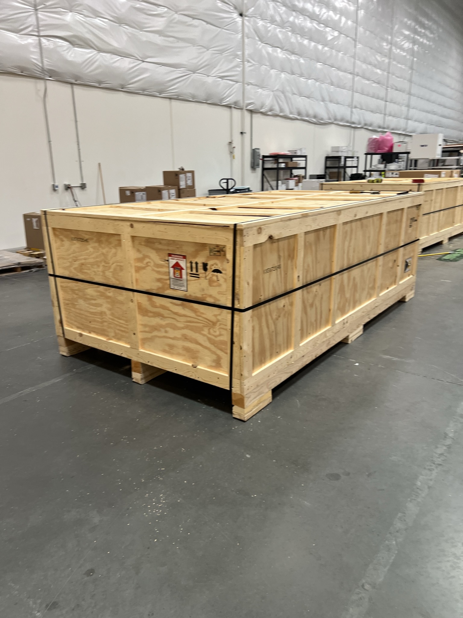 A very long, rectangular plywood shipping crate sitting on a warehouse floor. It is reinforced with black plastic banding and features a red "This Side Up" directional label along with standard handling icons.